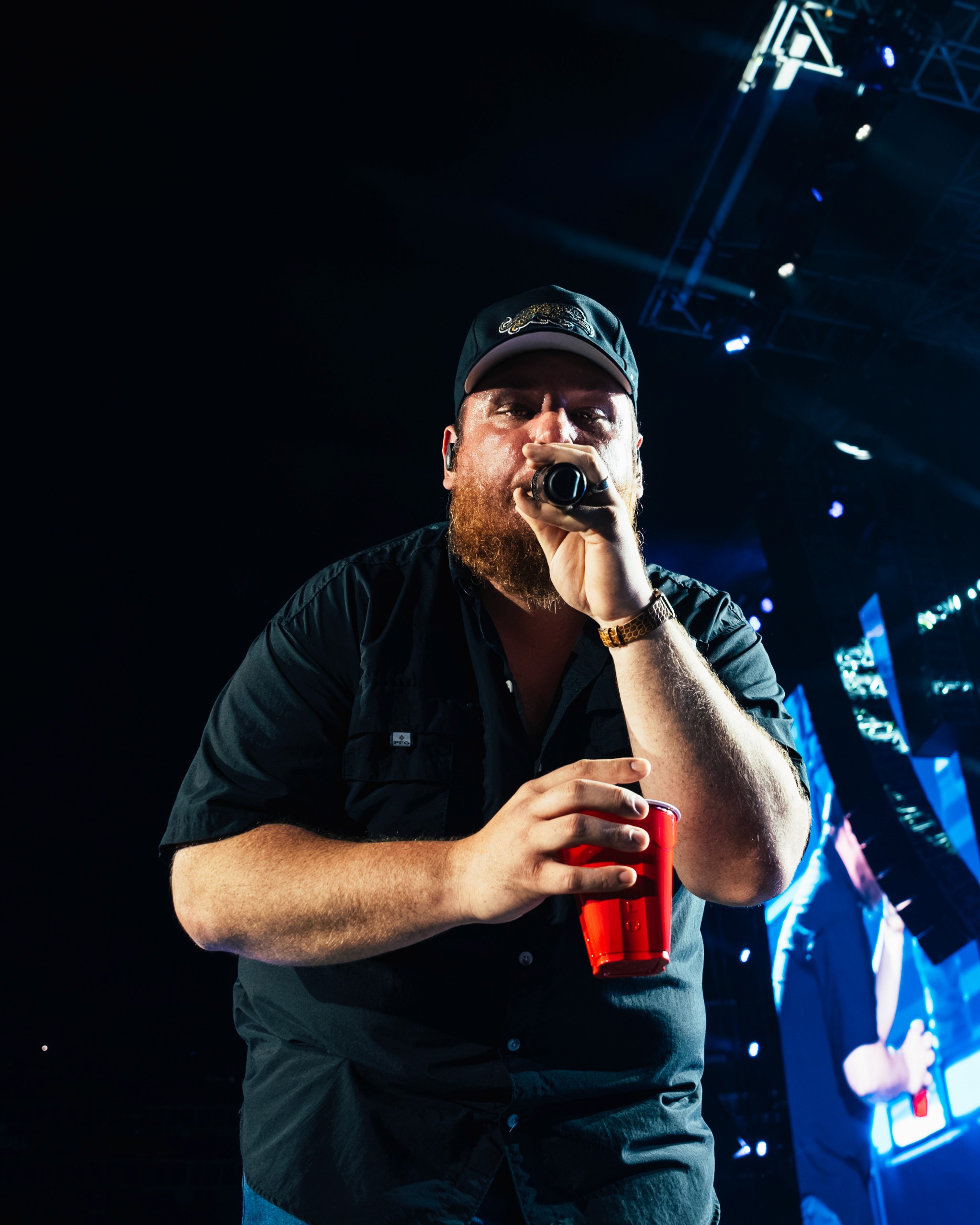 Luke Combs on the mic with a red Solo cup, the rig lit blue behind him.