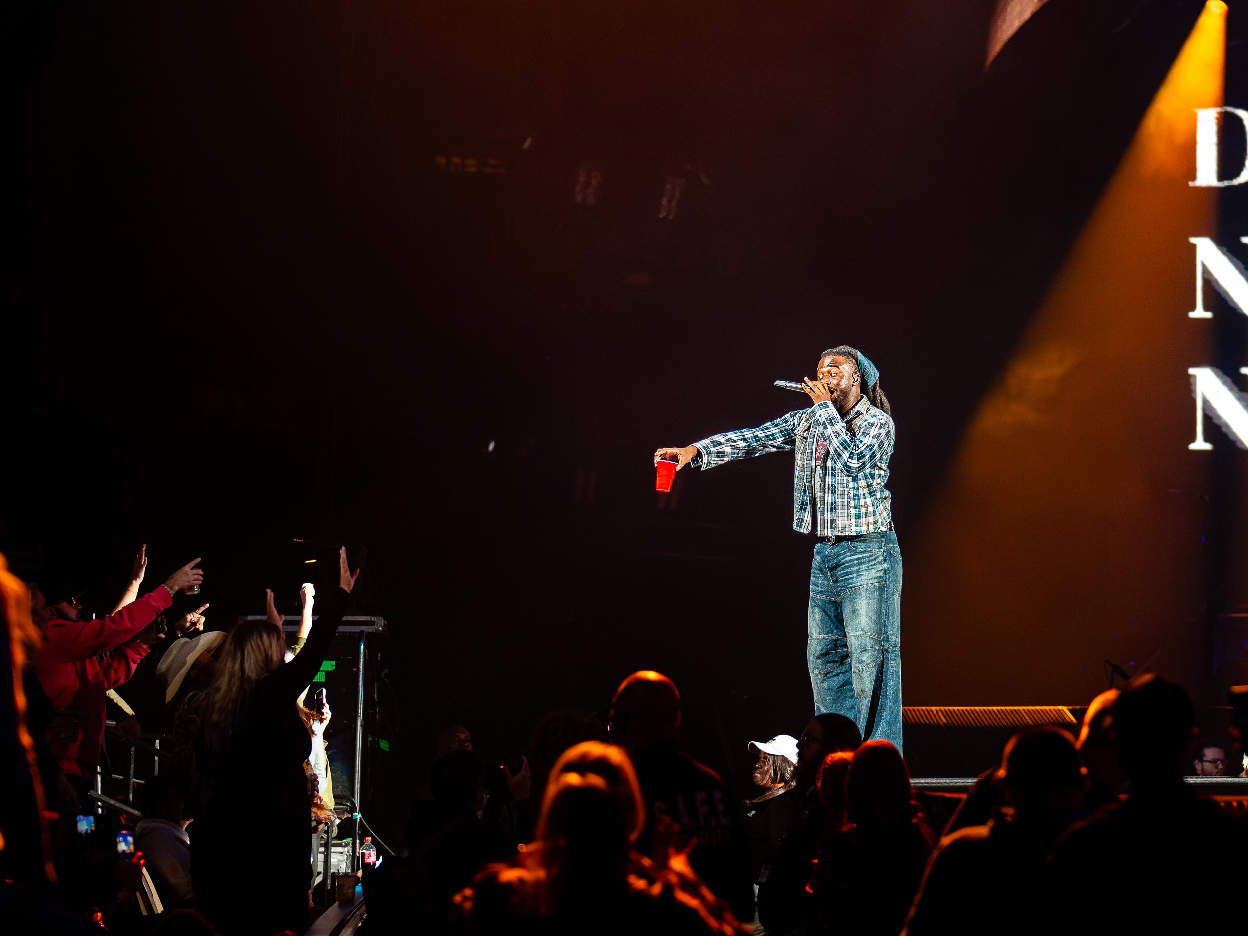 Shaboozey, red cup in hand, works the side of the stage with the front rows reaching.