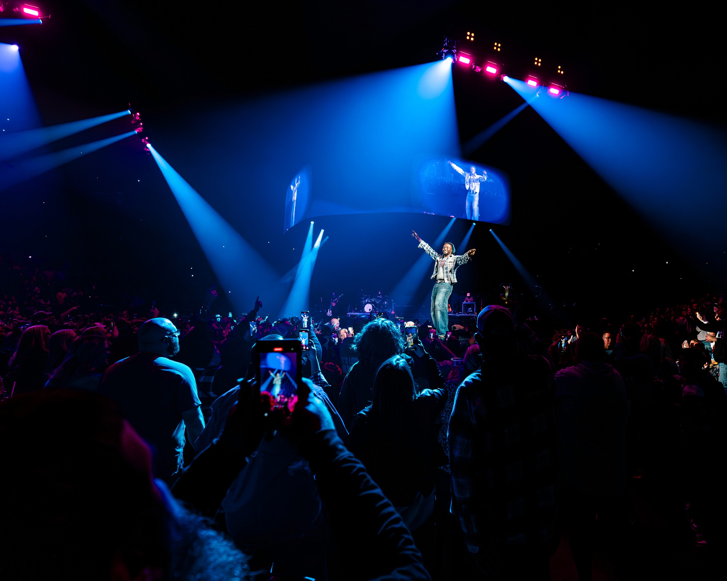 Shaboozey with both arms up on the runway, blue beams and his image on the screens, from the floor.