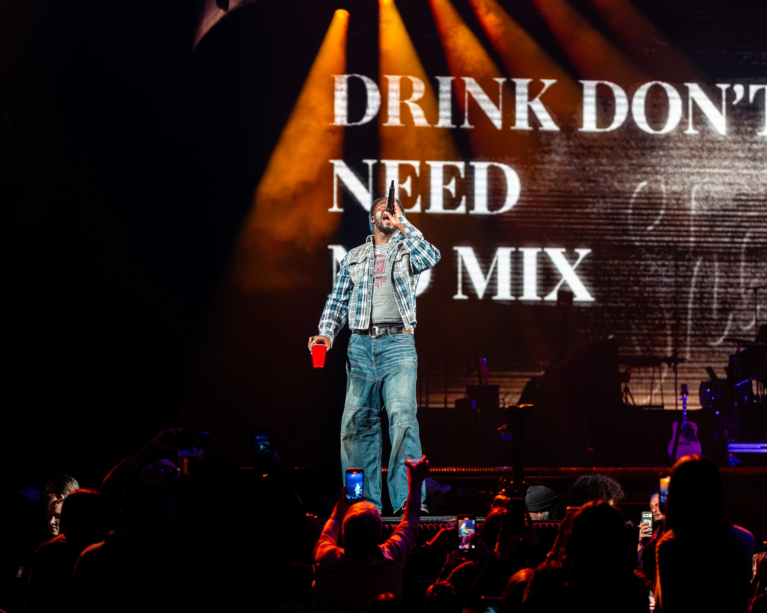 Shaboozey, cup in hand, under the words on the screen — DRINK DON’T NEED NO MIX.